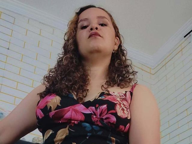 VittoriaI has curly brown hair, wearing a black floral dress with pink flowers, showing her tits, looking down at the camera with a slightly confident expression.