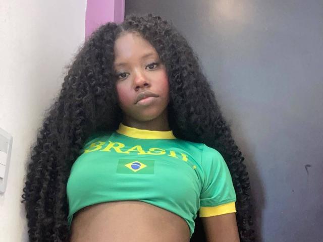 CatalinaSanA with long curly black hair, green Brazilian shirt, yellow trim, small tits, dark skin, pouty expression, standing against a dark wall, midriff exposed, looking at camera.