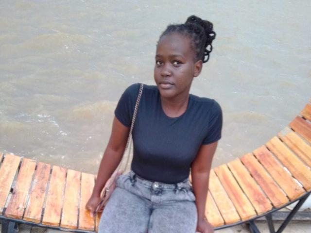 BlueBlossom, à la peau foncée et aux cheveux tressés en chignon, est assise sur un banc en bois, vêtue d'un t-shirt noir et d'un jean gris, et regarde légèrement sur le côté avec une expression neutre..