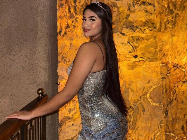 IsisFerrer with long black hair, wearing a sparkly silver dress, tiara, and smiling, standing with her back to the camera, looking over her shoulder at the camera.