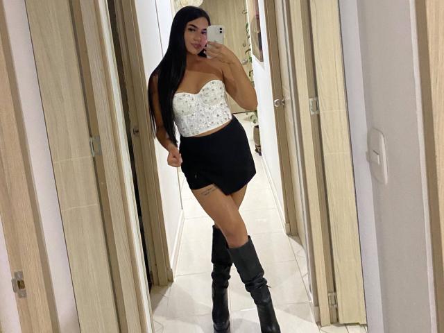 IsisFerrer with long black hair, white strapless floral top, black skirt, and knee-high black boots, taking a mirror selfie, showing her ass and thighs, in a hallway.