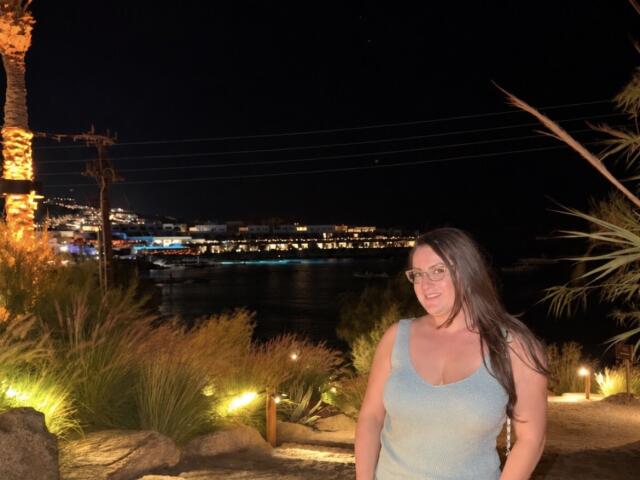 BadAngel with big tits in a blue tank top, smiling, has long dark hair, glasses, and is standing by a lit palm tree at night. Water and city lights in background.