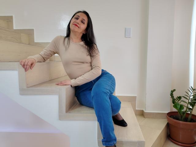 Cyntiia sitting on beige stairs, wearing a beige long-sleeve top, blue jeans, black flats, and red lipstick, with black hair and a slight smile. Her ass is on the stairs.