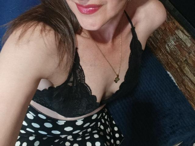 RubiAnderson with pink lips, black lace bra, and polka dot skirt, showing her tits and a gold necklace with a green star. Her arm is raised, smiling.