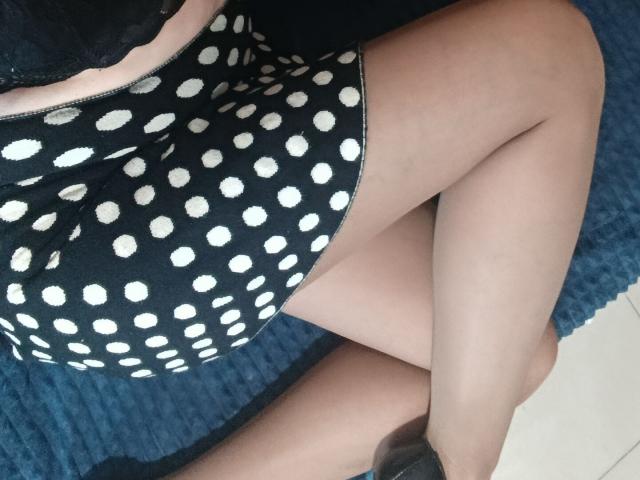 RubiAnderson's legs crossed, wearing a black polka dot dress, black lace top, and black heels, with pale skin, on a blue textured rug. Boobs visible under dress.