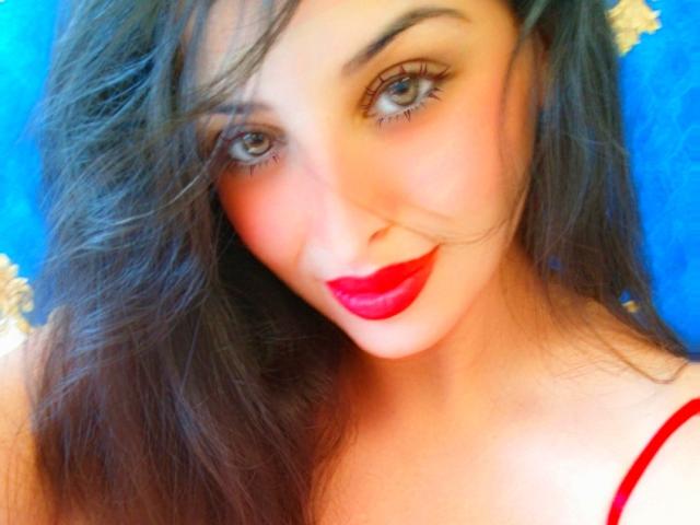 ScarletOra has red lipstick, blue eyes, and long brown hair, smiling slightly with a red strap visible on her shoulder. Her face is centered, looking directly at the camera.