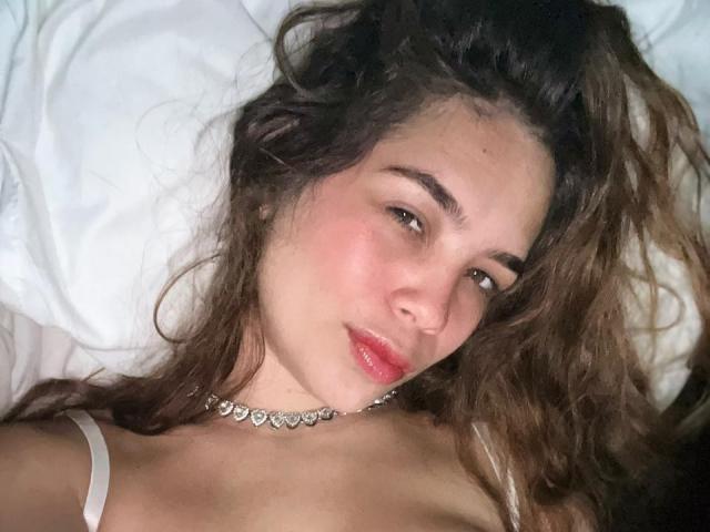 Cloe Santos, con i capelli castano ondulati, le guance rosate e le labbra rosse, indossa una collana d'argento, è sdraiata su cuscini bianchi, mostrando le spalle scoperte e lo spallino bianco..