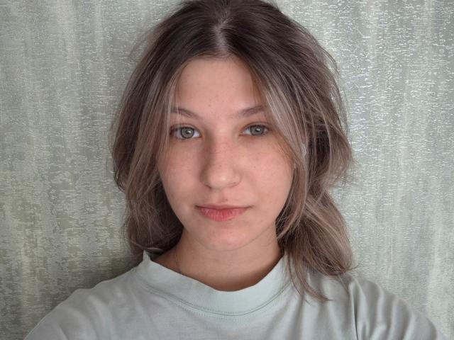 JennetteBuisson has light brown hair, green eyes, and a subtle smile, wearing a light gray shirt, with a textured gray background. Her face is slightly freckled.