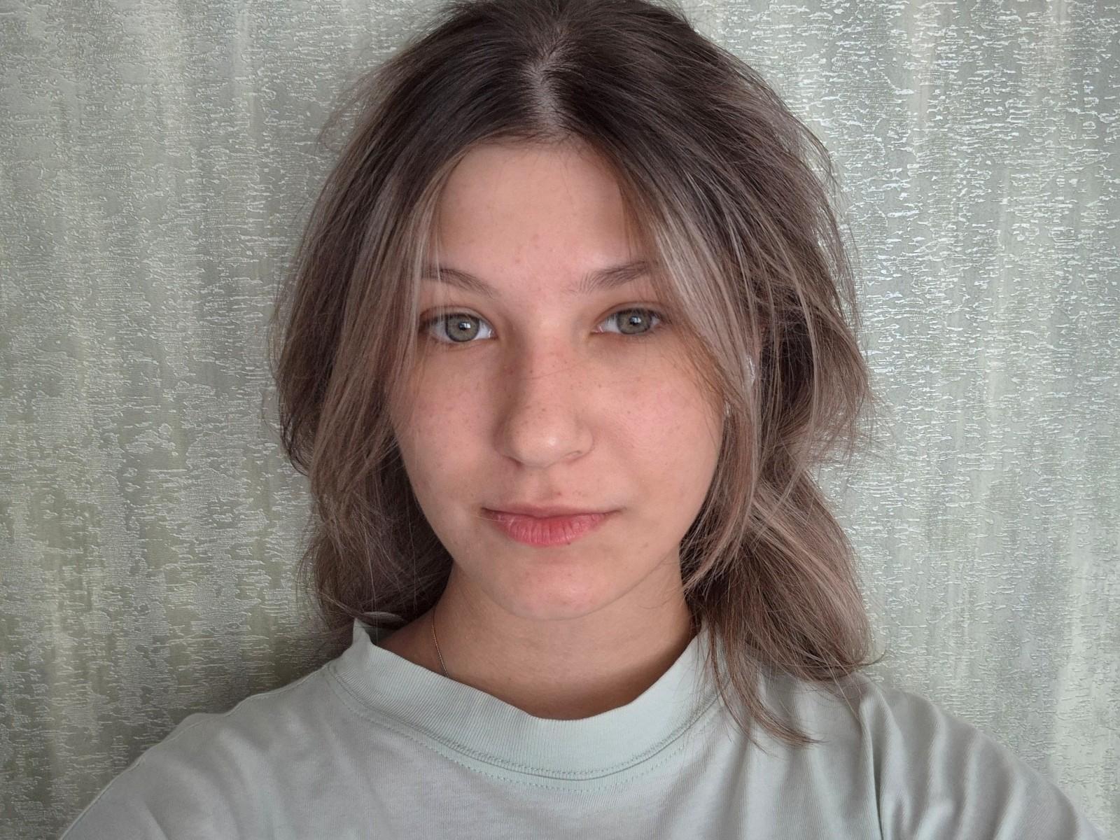 JennetteBuisson has light brown hair, green eyes, and a subtle smile, wearing a light gray shirt, with a textured gray background. Her face is slightly freckled.