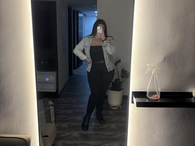 VenusxMusa, com cabelos longos e pretos, vestindo uma jaqueta jeans, um top preto e calças pretas, tirando uma selfie no espelho, mostrando seu traseiro com botas pretas de plataforma..
