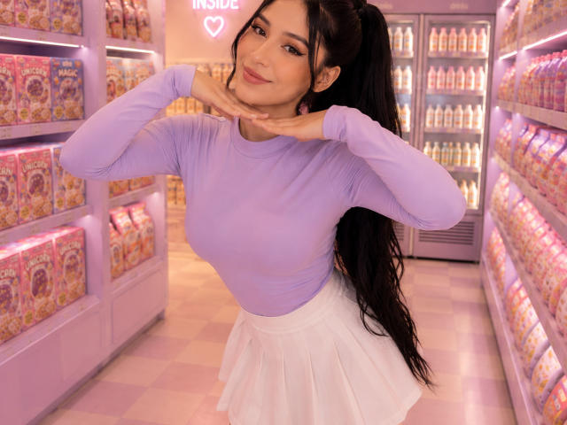 CelesteOcampo a de grandes seins, une longue queue de cheval noire, elle porte un haut violet à manches longues et une jupe blanche, elle sourit, les mains près de son visage, et se tient dans un magasin très éclairé..