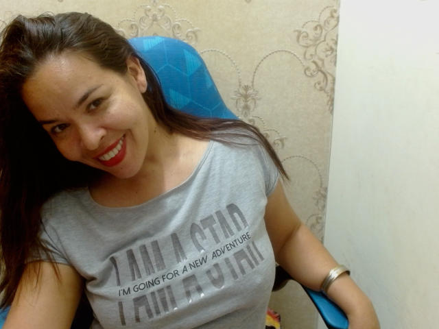 LizethBells, souriant avec du rouge à lèvres, a de longs cheveux bruns et porte un t-shirt gris, laissant légèrement entrevoir ses seins à travers le tissu, assise sur une chaise bleue..