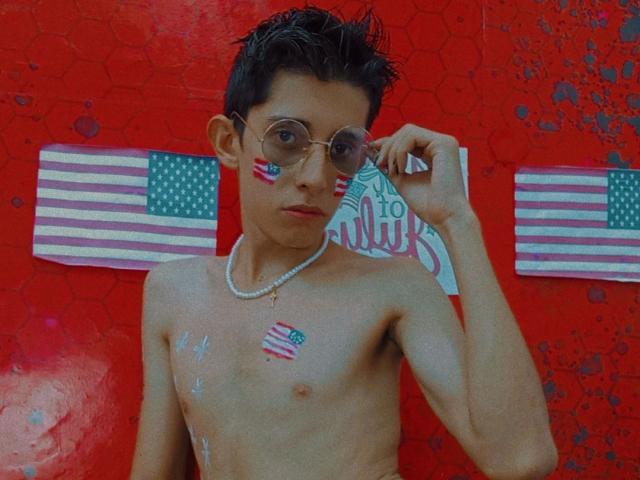 GomitaBay, con cabello corto y oscuro, usando gafas, un collar de perlas y pintura con la bandera estadounidense en la cara y el pecho, sin camisa, de pie frente a una pared roja con un patrón hexagonal..