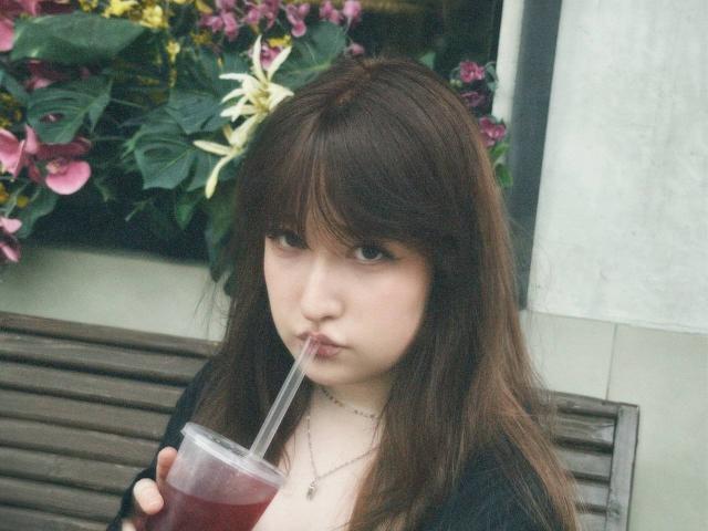 TrishaRidderhoff with brown hair, sucking a red drink through a straw, holding it in her right hand, wearing a black top, looking at the camera, on a wooden bench.