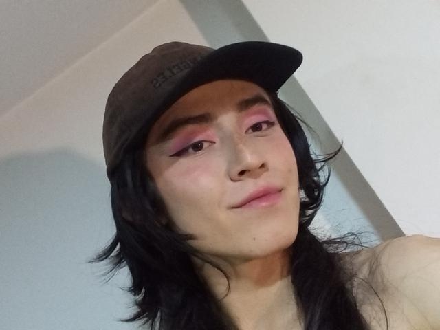 EllieCherry, avec ses cheveux noirs, son fard à paupières rose, sa casquette marron et son sourire discret, présente un visage mignon et une expression douce. Ses seins ne sont pas visibles, l'attention est portée sur le haut de son corps..