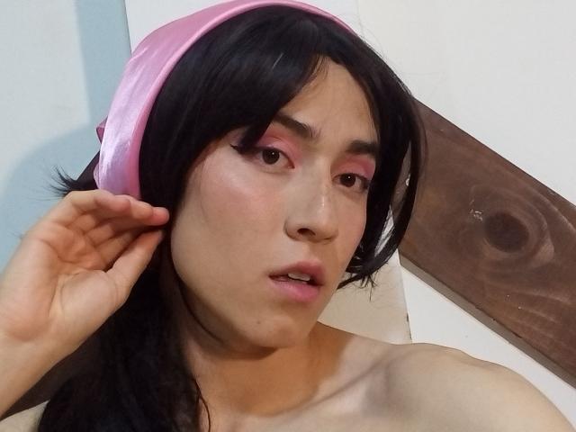 EllieCherry has black hair, pink eye makeup, and a pink velvet headband. She's topless, with her hand near her ear, and her mouth is slightly open.