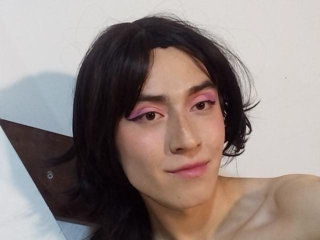EllieCherry has pink eyeliner, black shoulder-length hair, and a slight smile with visible collarbones. Their skin is light with a subtle freckle pattern. No clothes are shown.