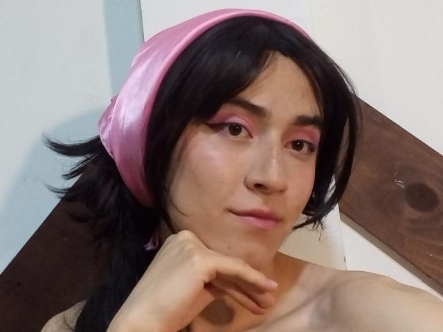 EllieCherry with black hair, pink headband, and pink eyeshadow, resting chin on hand, showing bare shoulders, subtle smile, and soft skin, looking at camera.