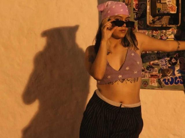 RubyLane, avec une poitrine de taille moyenne, portant un haut court violet avec des étoiles blanches, un pantalon à rayures, un bandeau rose et des lunettes de soleil noires, tenant un téléphone, avec une ombre sur un mur beige..