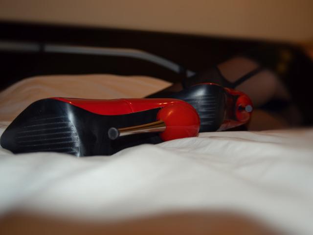Los elegantes zapatos de tacón rojo y negro de MistressOfShadow, con una punta de tacón plateada, descansan sobre una cama blanca. Las suelas tienen una textura, y los tacones están colocados uno al lado del otro..