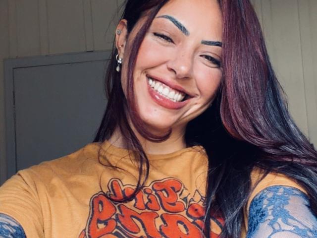 GaiaX, avec de longs cheveux roux-brun, souriante, montrant des dents blanches, portant un t-shirt jaune avec l'inscription "BBQ", et avec des tatouages bleus visibles sur les bras. Elle a des sourcils foncés et porte des boucles d'oreilles..