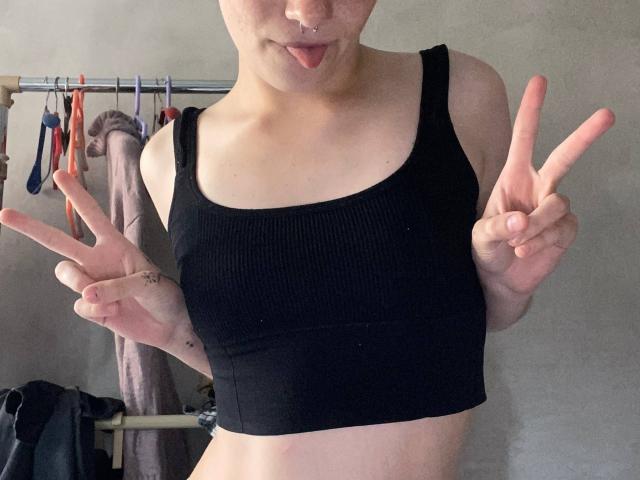 KarlaRolison with a nose ring, tongue out, wearing a black ribbed crop top, flashing dual double-v's, showing her small tits and a tattoo on her left hand.