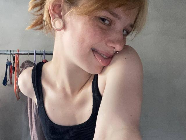 KarlaRolison with freckles, nose ring, and ear gauges, tongue out, black tank top, blonde hair, showing her shoulder, and a slight smile. Her tits are not visible.