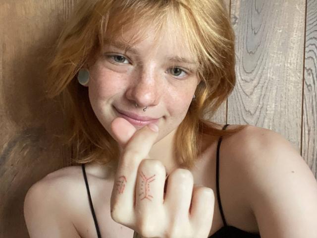 Karla Rolison mit roten Haaren, Nasenring und Ohrringen, lächelnd, zeigt einen Finger mit einem roten Symbol-Tattoo, trägt ein schwarzes Top mit Trägern..