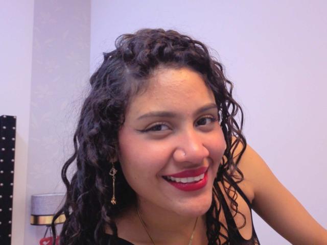 ValeryPoleth has curly black hair, red lipstick, and a nose ring, smiling with white eyeliner, wearing a gold necklace, and dangling earrings. She's looking slightly to the side.