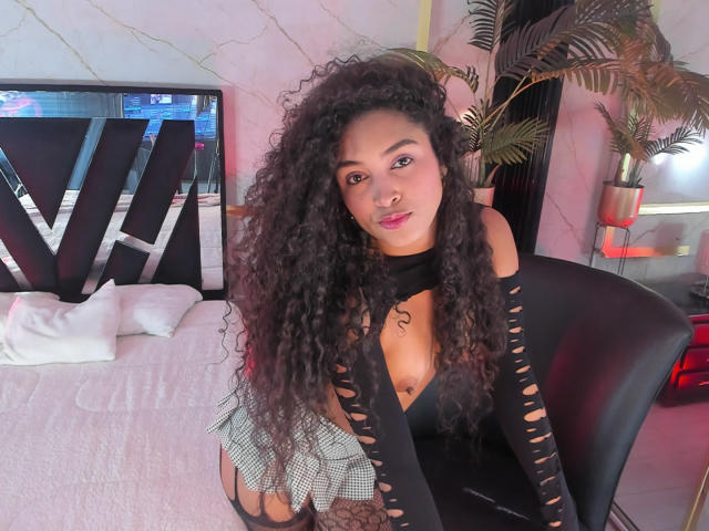 PaulinaRizzo with curly black hair, wearing a black cut-out top, exposing her tits, sitting on a black chair, looking at the camera with pink lipstick.