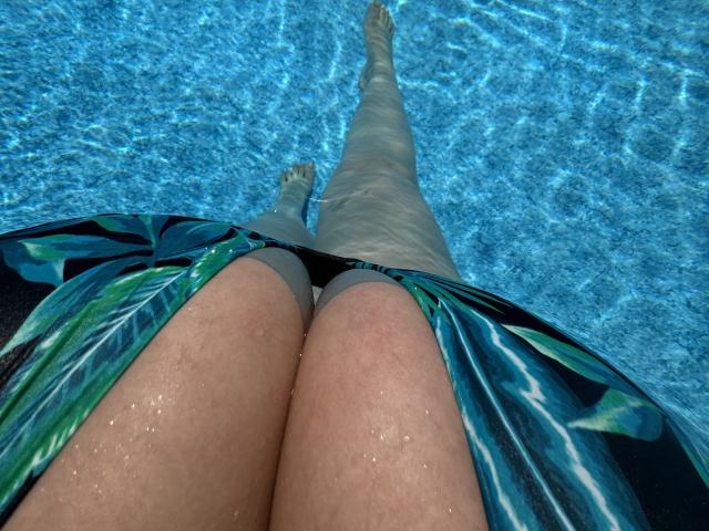 Barbarica's thighs in a blue-green floral bikini, water droplets on her skin, legs stretched out in a clear blue pool, taken from a downward angle.