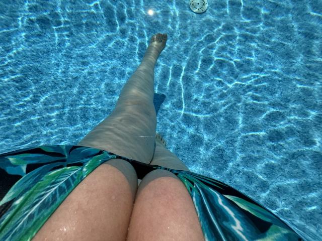 Barbarica's thighs and butt in a green leaf-patterned swimsuit, standing in a clear blue pool, with sunlight rippling through the water. Her legs are slightly apart, toes pointed.