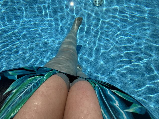 Barbarica's legs in a blue-tinted pool, wearing a green leaf-patterned dress, with her thighs and ass visible, and a metal pool ring above. Water ripples around.