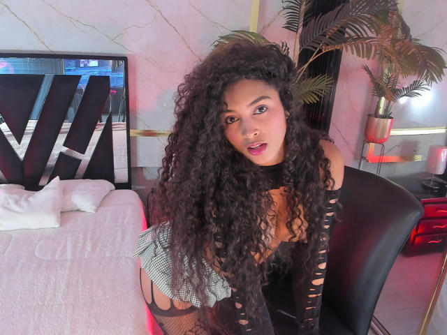 PaulinaRizzo with long curly black hair, wearing a ripped black top, green and white striped skirt, and black fishnet stockings, leaning forward with her tits and ass visible.