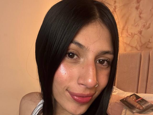 SophieBeaumont has shiny, slightly flushed skin, black straight hair, brown eyes, a nose ring, and a pink lipstick smile. Her tits are not visible. She's looking cute.