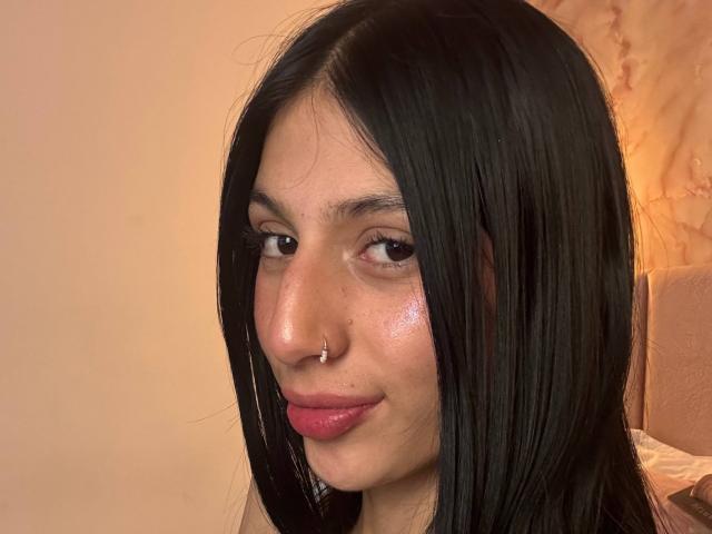 Sophie Beaumont a les cheveux noirs et lisses, un piercing au nez et du rouge à lèvres rose. Son visage brillant et son léger sourire sont visibles. Elle regarde l'appareil photo avec ses yeux marrons..