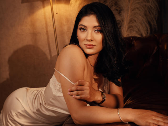 MaddieDemie with long black hair, wearing a white lace dress, lying on a brown leather couch, looking at the camera, her hand on her shoulder, warm lighting.