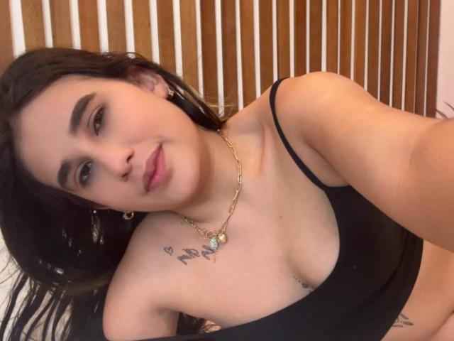 KatieDesirX, con i capelli neri, indossa una canotta nera, mostrando piccoli seni e un tatuaggio sopra il seno destro, sorridendo alla fotocamera. Indossa una collana d'oro..