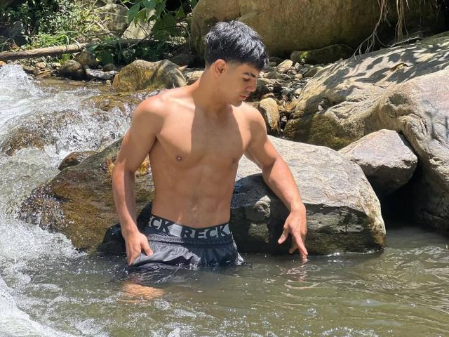 MasonHoney, torse nu, avec une musculature tonique, se tient dans un ruisseau rocheux, l'eau montant jusqu'à son short noir RICKERDICKER, et regarde vers le bas avec un léger sourire, ses cheveux noirs courts..