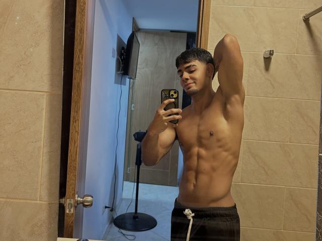 MasonHoney, sin camisa, con un torso musculoso y definido, pantalones cortos negros y una sonrisa, se toma un selfie frente a un espejo, mostrando sus abdominales, en un baño. Está sosteniendo un teléfono..