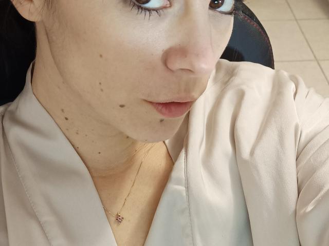 OrianaLaFrancaise has light skin with freckles, wearing a beige shirt, and a gold necklace with a pink gem. Her face is slightly turned with a pouty expression.