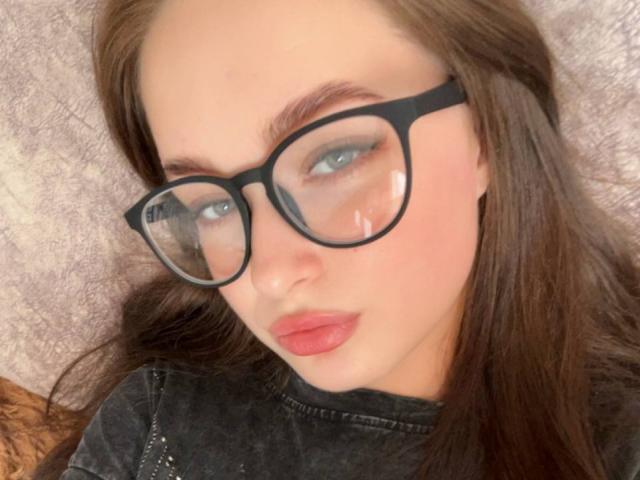 Melania Rannels, avec de grandes lunettes à monture noire, des yeux bleus et du rouge à lèvres rose, a un visage sérieux et de longs cheveux bruns, et porte un haut noir, sur une photo de profil légèrement floue..
