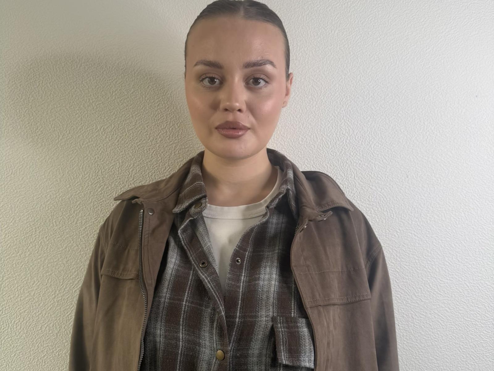 Olivia Azbill tiene la cabeza rapada, ojos marrones y lleva una chaqueta marrón sobre una camisa de cuadros con una camiseta blanca. Sus glúteos no son visibles..