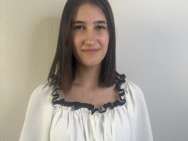 NellValentia with straight brown hair, small tits, wearing a white blouse with black ruffled trim, smiling softly, standing against a textured white wall.