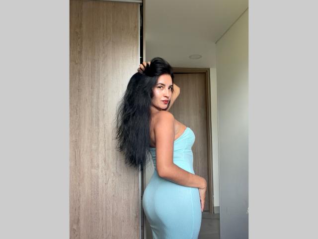 Emily Jensenn, con cabello negro largo, vestido azul sin tirantes y trasero curvilíneo, de pie en un pasillo, mirando hacia atrás, con la mano en el pelo, y una puerta de madera en el fondo..
