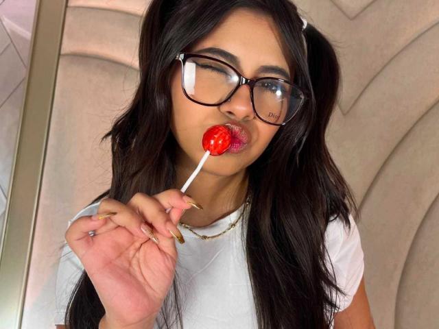 CelesteMorel with long dark hair in pigtails, wearing glasses, sucking on a red lollipop, winking, white shirt, gold necklace, and gold rings, in a selfie.