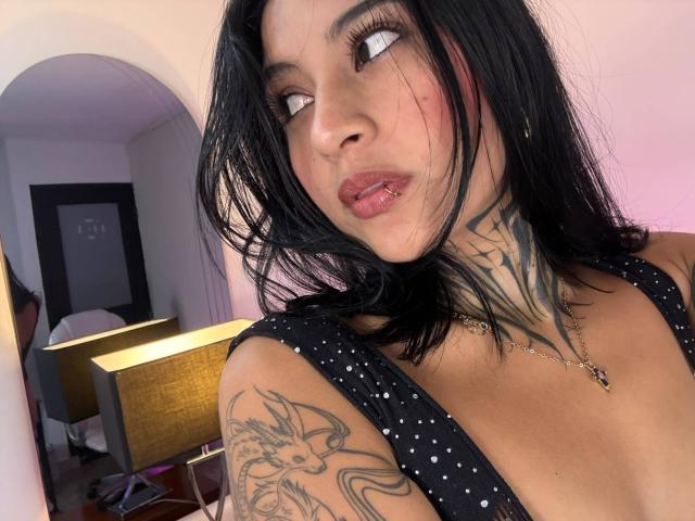 NicolleSweetty heeft zwart haar, tatoeages en draagt een zwart topje met stippen. Ze kijkt opzij, met haar lippen lichtjes open, waardoor haar roze lippenstift zichtbaar is..