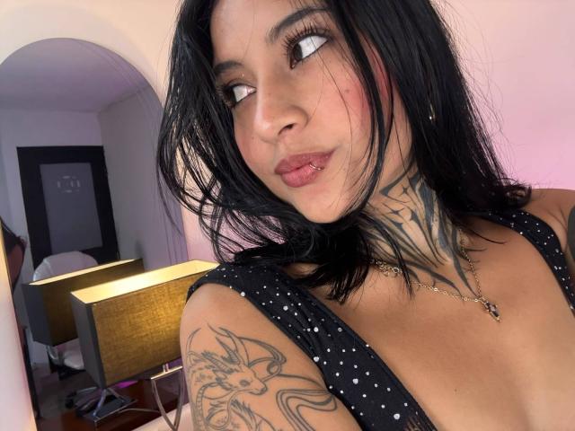NicolleSweetty with black hair, white eyeliner, and pouty lips, wearing a black polka dot top, showing tattoos on her neck and arm, and a gold necklace.