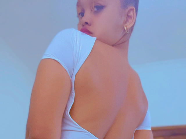 EloCherie est dos à la caméra, on voit son cul nu et un haut blanc décolleté, avec une vue de profil de son visage et une boucle d'oreille dorée..