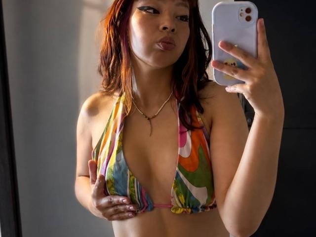 Viky Evans, con i capelli rossi, indossa un top di bikini colorato e a motivi, sta scattando un selfie allo specchio, tenendosi il seno con una mano, con un'espressione accigliata, e tiene in mano un telefono bianco..